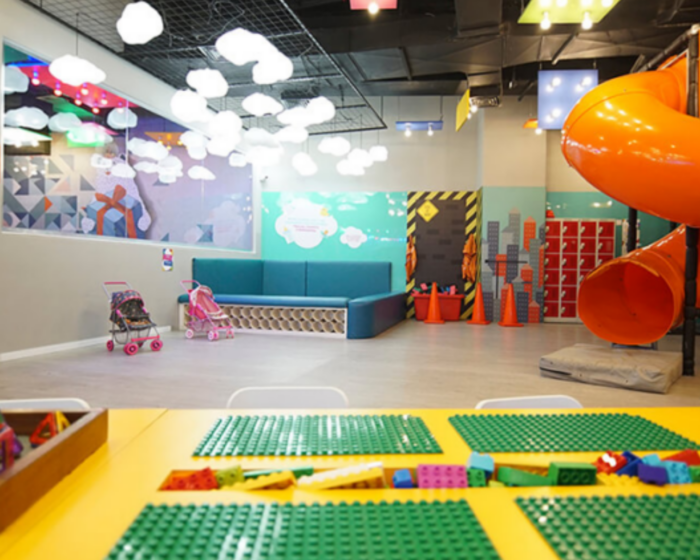 Foto de um ambiente interno decorado com muitas cores. Há um escorregador em tubo curvo, uma mesa de brincar e sofás com brinquedos,