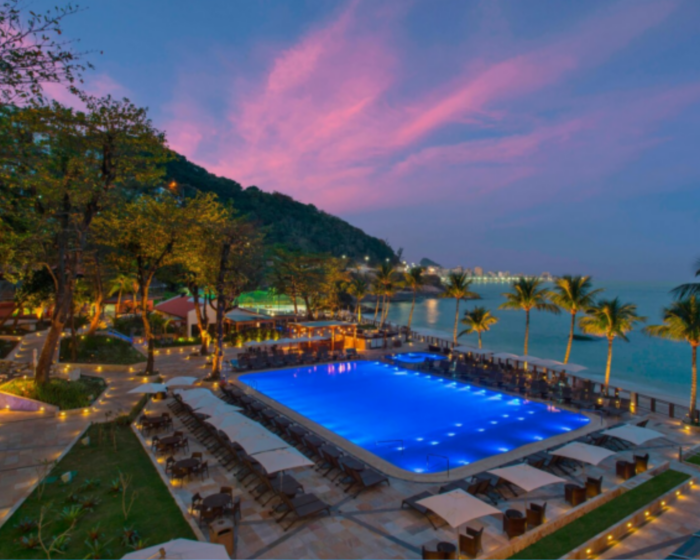 Vista da piscina do hotel Sheraton iluminada ao pôr do sol; Ao fundo, palmeiras, espreguiçadeiras, o mar e uma montanha