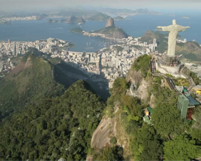 Foto da vista aérea do Cristo Redentor com o horizonte do Rio de Janeiro ao fundo, aparecendo a Baía de Guanabara, morros e prédios da cidade. O Cristo Redentor é uma estátua cinza clara enorme com os dois braços abertos.