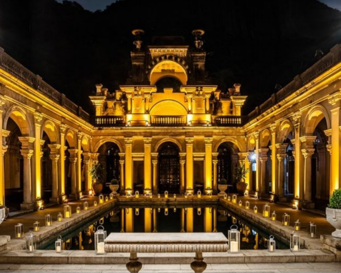 Foto do Parque Lage à noite. Ao centro, uma piscina iluminada pelas luzes da arquitetura ao redor, com vários pilares formando uma construção em formato de um U horizontal. À frente da piscina, um banquinho branco.
