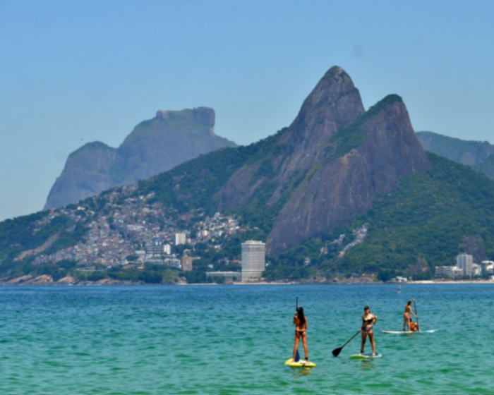 Foto da praia do Arpoador na qual aparece apenas a água do mar e os morros cariocas ao fundo. Na água cristalina e clara, três pessoas fazendo stand up paddle.