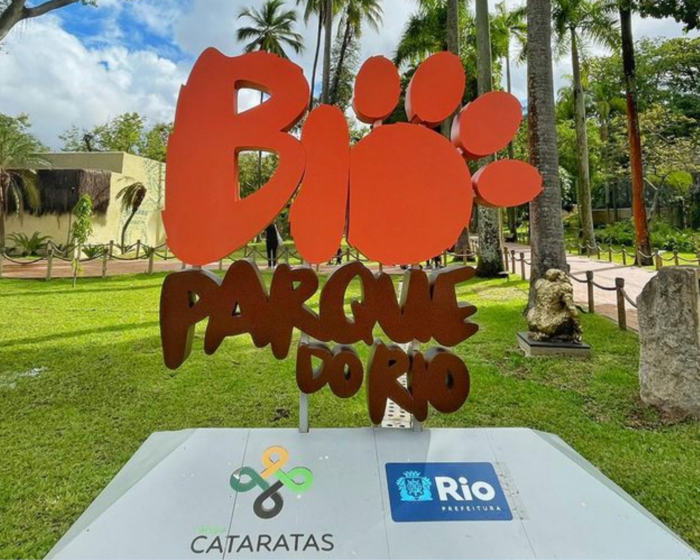 foto de uma placa em 3D e tamanho grande com a logomarca do BioParque do Rio. Nela, o O de Bio ganha dedinhos e vira uma pata animal. Ao redor, a paisagem do zoológico com grama, árvores e céu azul com nuvens brancas.