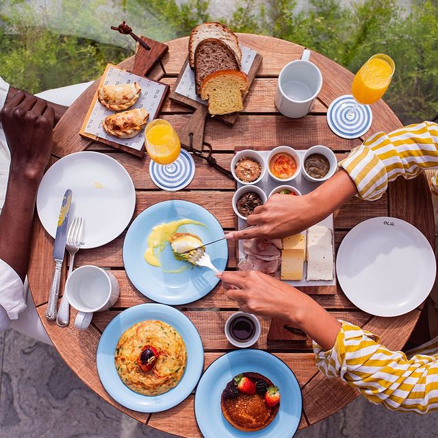 Foto aérea de uma mesa redonda de madeira com várias comidas diferentes espalhadas. Em volta da mesa, duas pessoas que não aparecem por completo na foto, e sim omo braços. Uma delas é branca e a outra, negra.