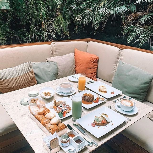 Foto de perto de uma mesa quadrada de madeira posta com vários pratos de comida em cima. Ao redor, um sofá de canto com almofadas e uma parede de plantas.