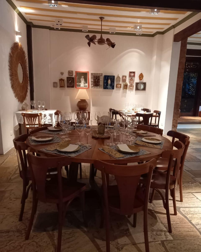 Foto de um ambiente interno de restaurante. Uma mesa redonda de muitos lugares está posta com pratos, copos, talheres e decoração. As cadeiras também são de madeira. As paredes próximas são branas e decoradas com objetos na mesma paleta de cores.