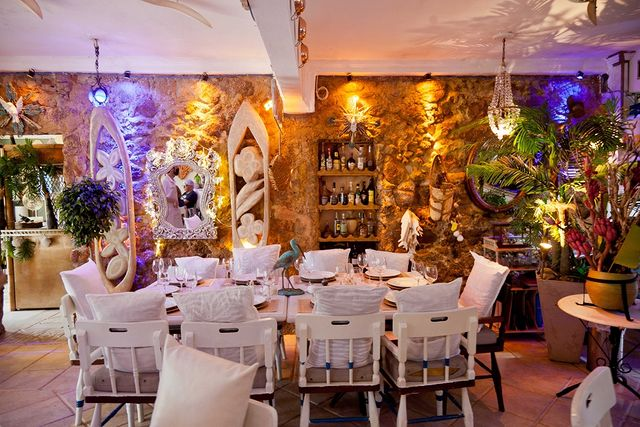 Foto de interior de restaurante. Mesa branca retangular e comprida está posta, cercada por cadeiras que combinam. A iluminação é com spots apontados para as paredes e refletindo na textura delas. Há plantas complementando a decoração.