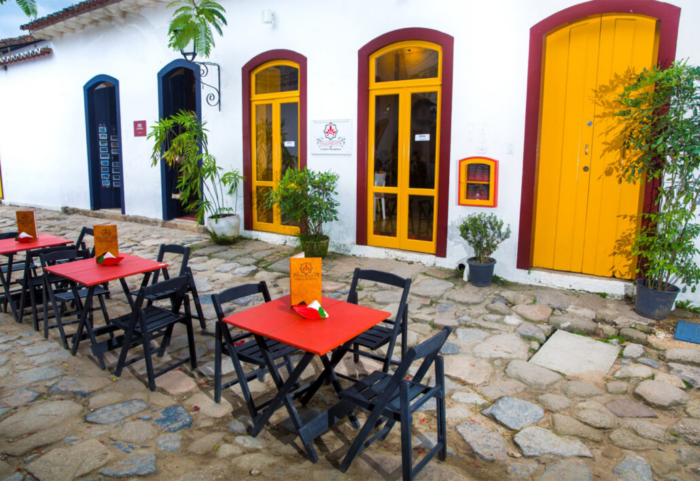 Foto de um exterior de restaurante. Mesas pequenas de até quatro pessoas estão colocadas na calçada, forradas com toalhas de mesa e decoradas com centros de mesa. As cadeiras ao redor são azuis. Ao fundo, a fachada do restaurante, uma parede branca com três portas imponentes amarelas.