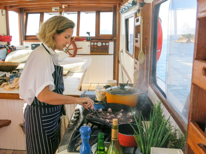 Foto de uma mulher cozinhando. Ela está de lado para a câmera e de frente para um fogão dentro de um barco. Nas mãos, uma frigideira com camarões e uma colher de pau mexendo. Ao fundo, o interior do barco.