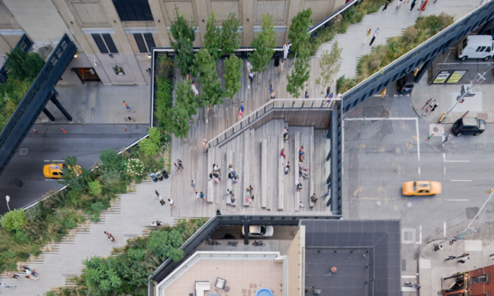 Vista aérea do High Line Park de Nova York. Um viaduto de madeira por cima de uma rua da cidade, com plantas, bancos de madeira e pessoas caminhando.