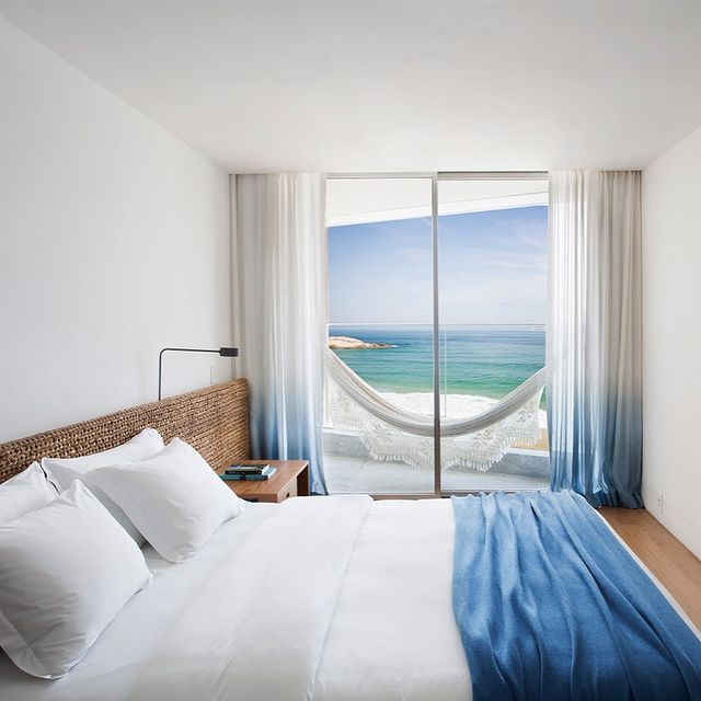 Foto de um quarto de hotel com paredes brancas, cortinas brancas abertas revelando uma janela do chão ao teto. A vista da janela é de uma rede branca pendurada na varanda e, ao fundo, o mar. A cama do quarto é grande e está feita com jogo de cama branco com detalhes azuis no pé.