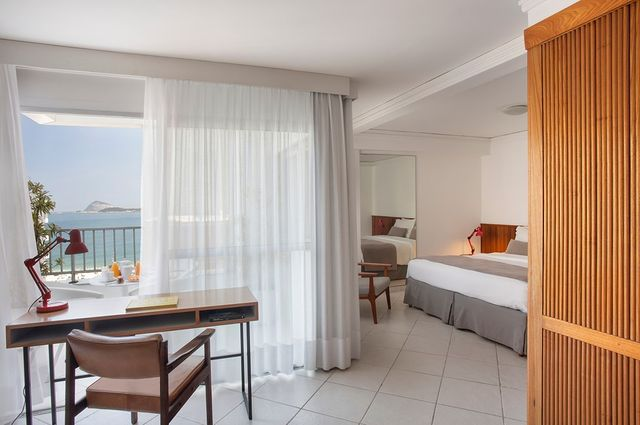 Foto de um quarto de hotel espaçoso. Em primeiro plano está uma mesa de madeira com cadeira acolchoada, luminária e logo em frente a varanda, coberta por uma cortina de pano leve branco. Na vista, o mar. À direita, o quarto se abre na área da cama e do armário, com um espelho grande.