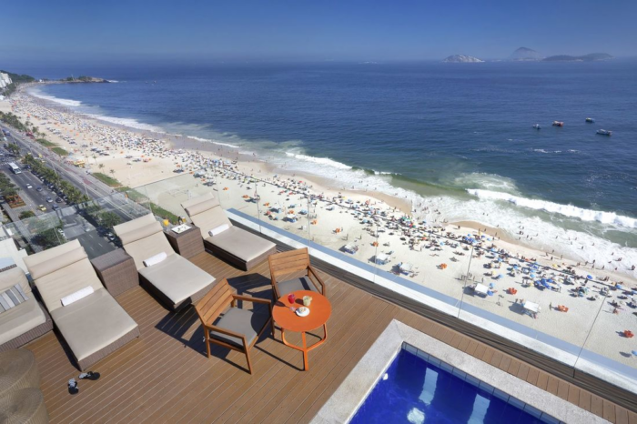 Foto aérea da vista da cobertura do hotel, mostrando a praia à distância, espreguiçadeiras, um deck de madeira e um pedaço da piscina