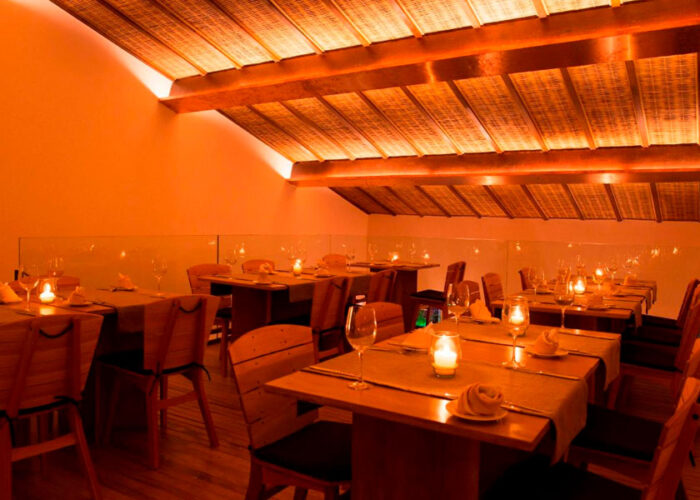 Foto de um interior de restaurante. A iluminação é alaranjada e o teto é curvo, mais baixo nos cantos. As mesas são retangulares de madeira e estão postas com pratos, talheres e copos.