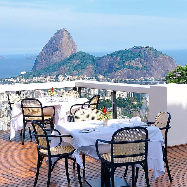 Foto de um ambiente aberto de restaurante, em uma varanda. As mesas são quadradas e estão com toalhas de mesa brancas, cada uma posta com talheres, copos e um pequeno vaso cilíndrico com flor dentro. Ao fundo, o Pão de Açúcar e uma parte do horizonte do Rio de Janeiro.